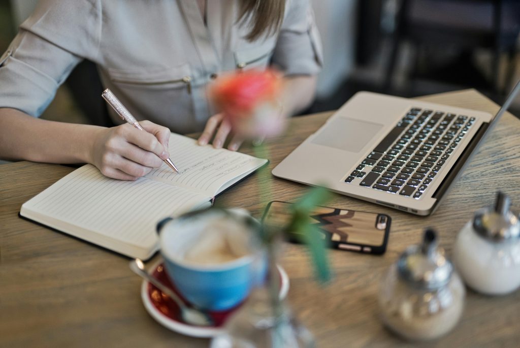 Woman writing in a notebook with a laptop and coffee cup on a desk. Ideal for workspace inspiration.